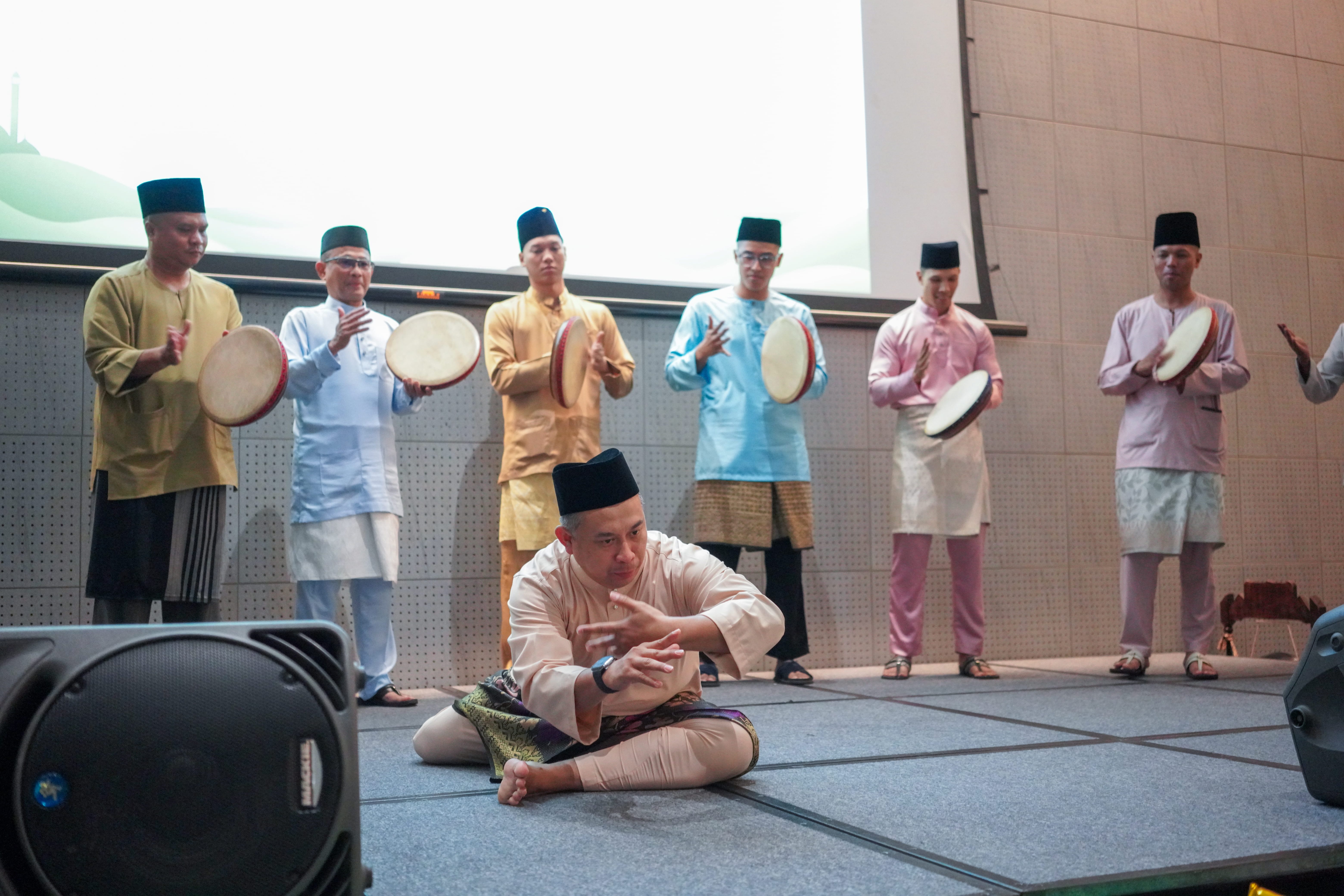 A silat performer demonstrating a seated martial arts pose on stage while kompang musicians play frame drums behind him.